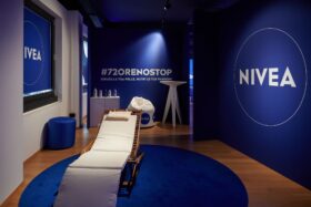 "Spazio relax NIVEA Body con lettino e decorazioni blu per l'evento #72ORENOSTOP, dedicato all'idratazione della pelle e al benessere personale."